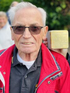 Ein älterer Mann mit Sonnenbrille schaut in die Kamera, im Hintergrund hält jemand einen Stolperstein hoch.