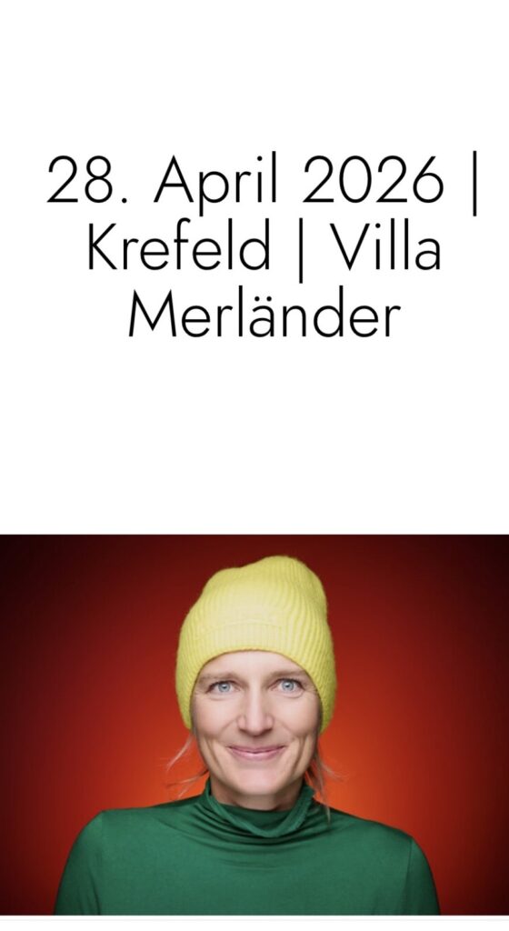 Eine Frau mit gelber Mütze und grünem Pullover steht vor einer roten wand und lächelt in die Kamera.