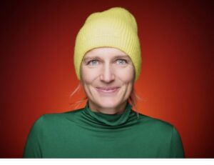 Eine Frau mit gelber Mütze und grünem Pullover steht vor einer roten wand und lächelt in die Kamera.