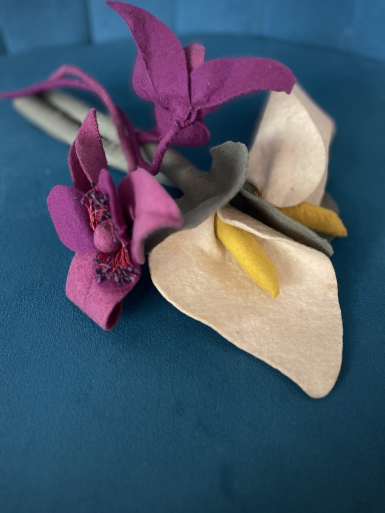 zwei Filzblumen, eine weiss-gelb, eine lila, liegen auf einem petrol blauen Hintergrund