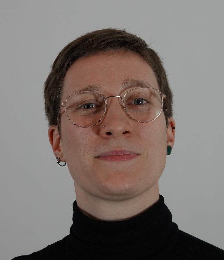 Portrait einer frau mit kurzen Haaren und Brille. Sie trägt einen schwarzen Rollkragenpullover und steht vor einer weißen wand.