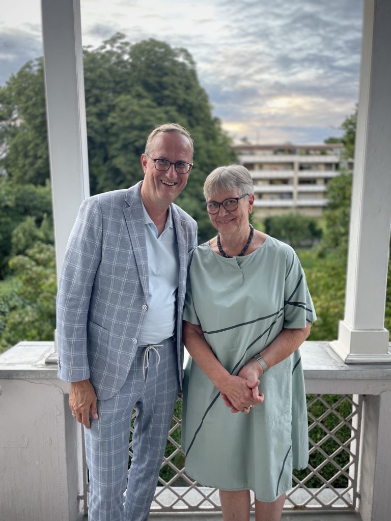 Ein Mann und eine frau (von links nach rechts) stehen im Freien vor zwei Säulen, im Hintergrund ist Gras und ein Haus zu sehen. Sie lächeln beide in die Kamera