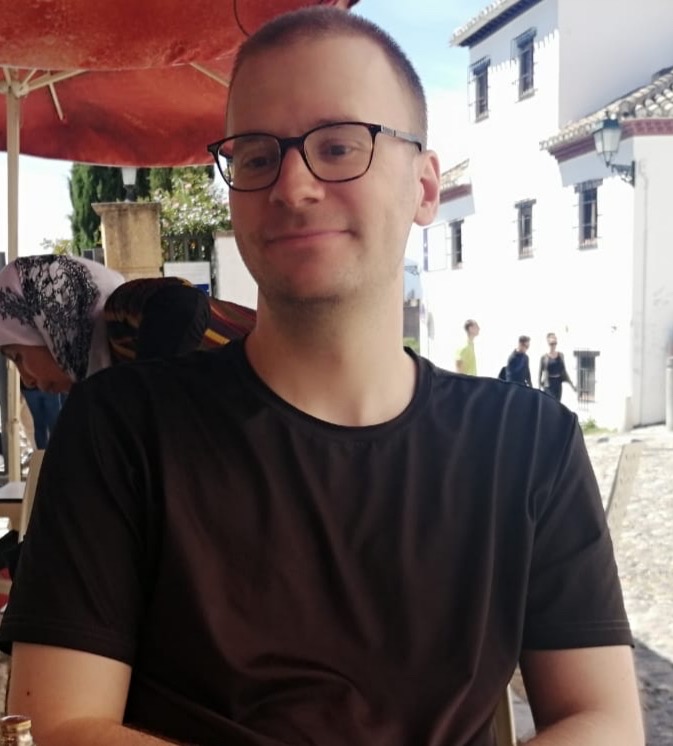 Ein junger Mann lächelt in die Kamera, er trägt eine Brille und ein schwarzes T-Shirt, im Hintergrund ist ein roter Sonnenschirm zu sehen