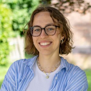 Eine junge Frau mit Brille und blonden Locken lächelt in die Kamera. Sie trägt eine blaue Bluse und steht draußen in der Natur
